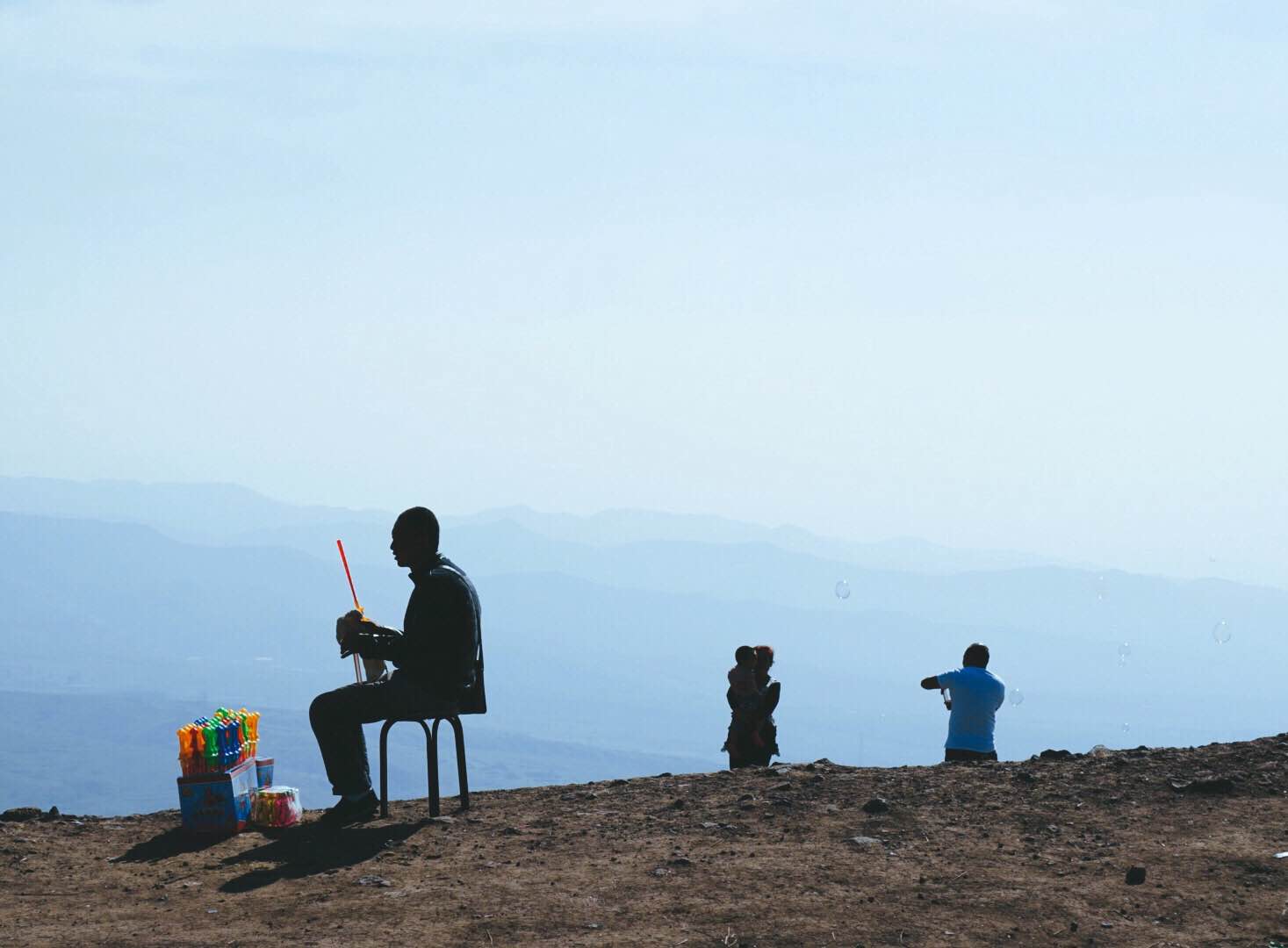 This is a photographic image of a man seated atop an unidentified mountain location. He is selling plastic bubble blowing tubes to children. Against the blue of the sky and vast landscape in the distance, the lone figure is symbolic of the contingency of our civic life in the expanse of planetary environment.